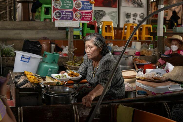 Elderly woman cooking traditional Thai dishes at a bustling Bangkok street market.