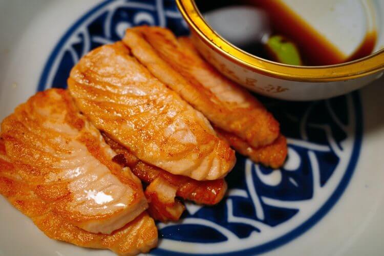 a blue and white plate topped with sliced fish next to a bowl of sauce