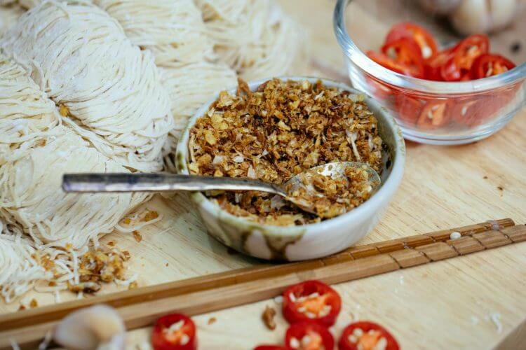 From above bowl of spicy condiment placed on wooden board near uncooked homemade noodles chopsticks and cut hot red peppers