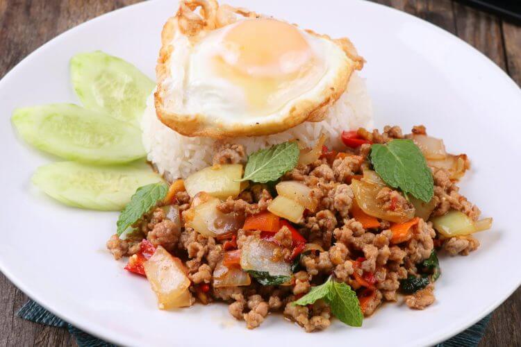 Authentic Thai basil pork with rice, fried egg, and garnished cucumbers on a white plate.