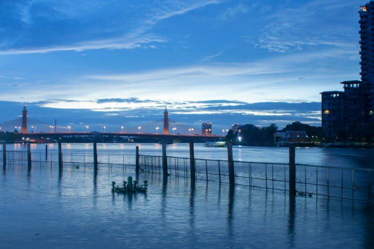 Scenic view of Wat Bowon Niwet, Bangkok, featuring an illuminated bridge over the water during twilight.