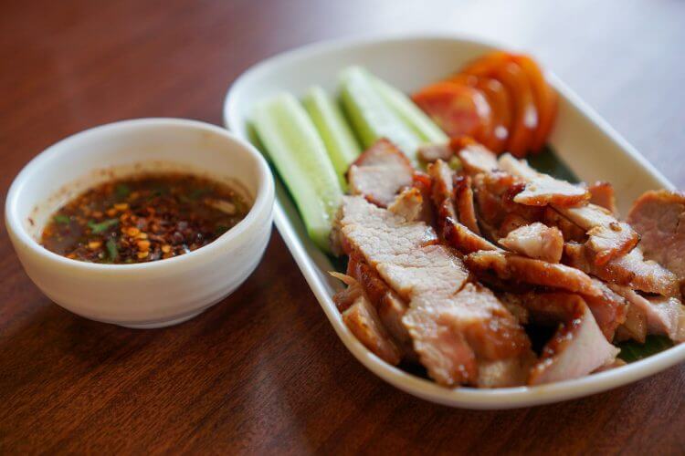 Close-up of grilled pork slices with cucumber, tomato, and dipping sauce for a flavorful meal.