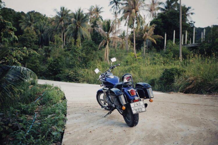A blue motorcycle parked on a sandy road surrounded by lush, tropical vegetation and palm trees.