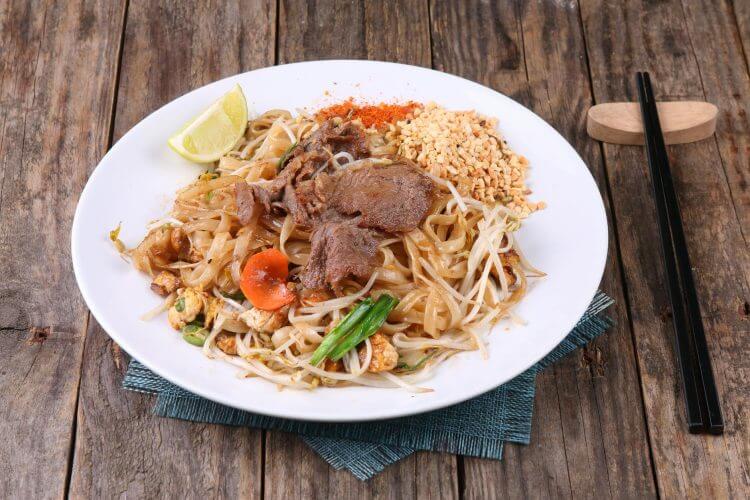 A vibrant beef Pad Thai dish served with lime, peanuts, and chopsticks on a rustic table.