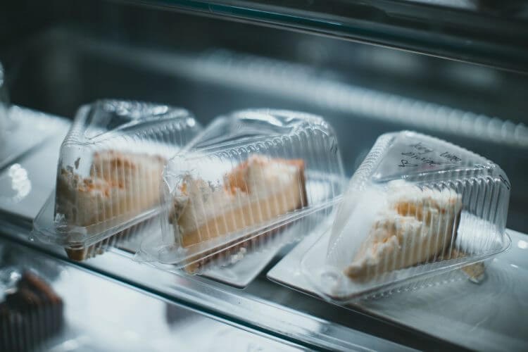 Three pieces of cheesecake in plastic containers are displayed in a refrigerated shelf.